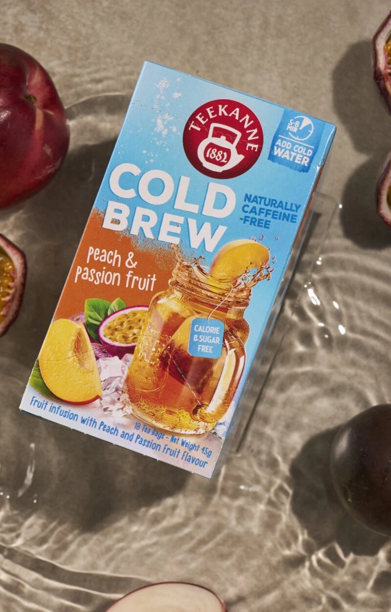 On the image you can see a COLD BREW tea box of the flavour peach-passionfruit. It is lying in a paddle of water in the sand surrounded by fresh passionfruit